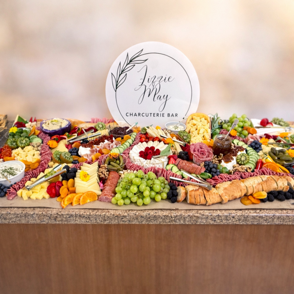 Charcuterie board with various meats, cheeses, fruits, and vegetables on a wooden surface with 'Lizzie May Charcuterie Bar' sign.